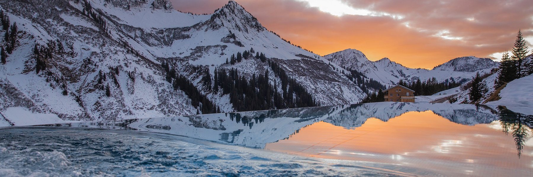 Infinity Pool mit Spiegelung der Bergkette im Winter bei Abenddämmerung im Panoramahotel Alpenstern