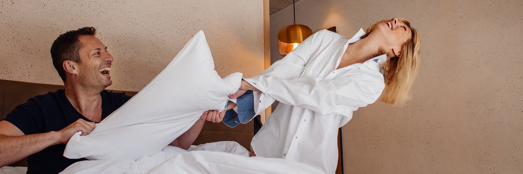 Mann und Frau lachen in einem Hotelbett und streiten sich um einen Kopfpolster
