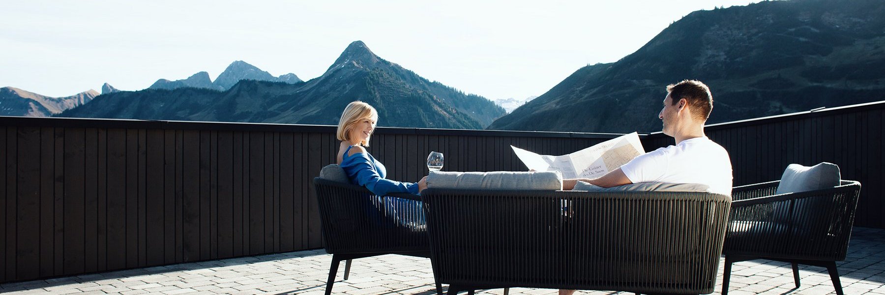 Frau und Mann auf der Terrasse mit Champagner und Zeitung mit Blick auf das Bergpanorama in Damüls